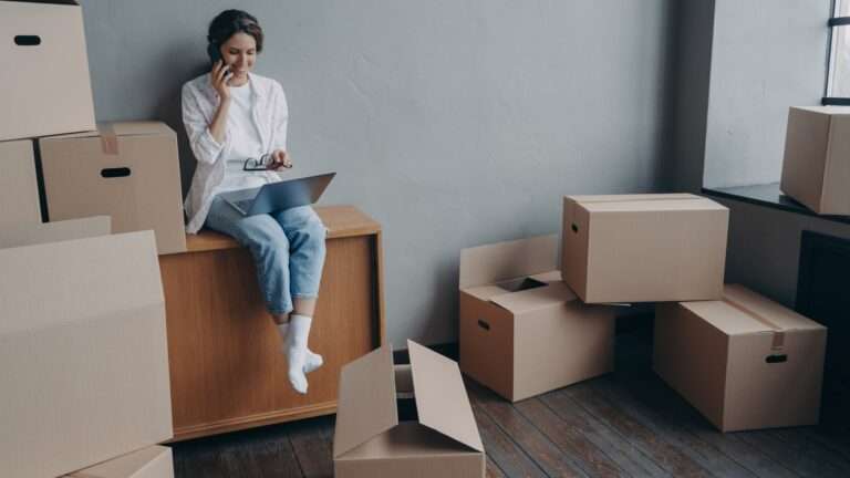 Happy homeowner talking on phone hispanic girl is sitting among carton boxes and working on pc